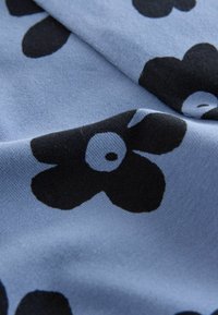Ljusblått tyg med stora svarta blommönster med cirkulära vita mittpunkter, som visar upp en slät textur och mjuk drapering.