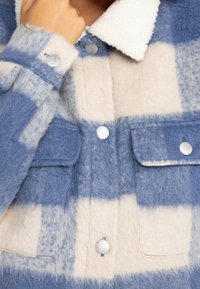 Nahaufnahme einer Person, die eine blau-beige karierte Wolljacke mit silbernen Knöpfen und aufgesetzten Taschen trägt, die Hand in der Nähe des Kragens.