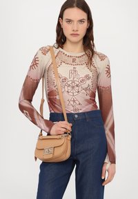 Langarm, hellbraunes Oberteil mit grafischen Mustern und einer beigen Umhängetasche mit einem silbernen Logo-Verschluss. Dunkelblaue hochgeschnittene Jeans.