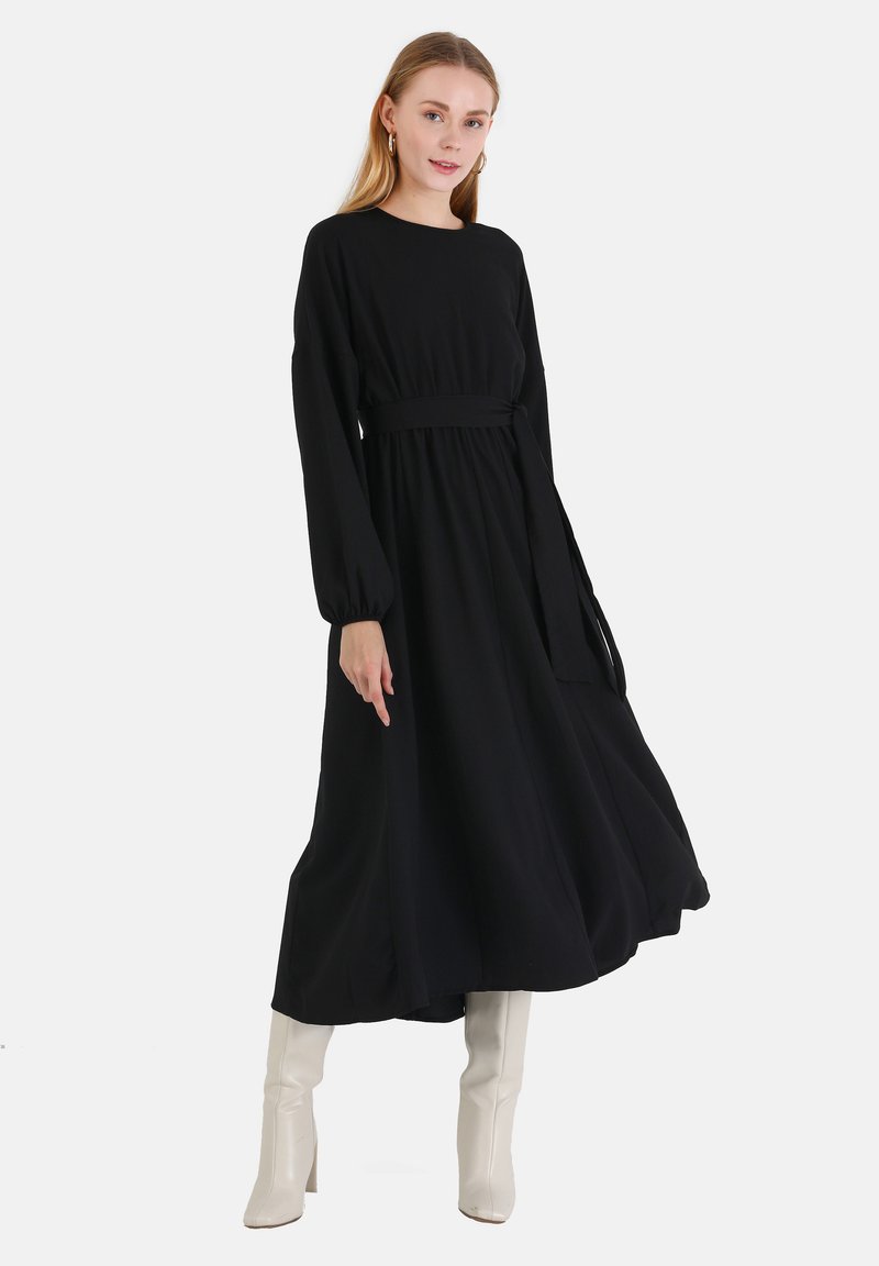 Schwarzes langärmliges Kleid mit einem Taillenbund, fließender Silhouette und weicher Textur. Kombiniert mit weißen kniehohen Stiefeln.