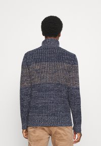 Pull en maille marine et beige avec un col haut, présentant un motif texturé et une bordure côtelée. Le design comprend un dégradé de couleurs.