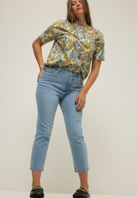 Camicia a maniche corte con motivo floreale gialla e blu, abbinata a jeans dritti blu chiaro e scarpe slip-on nere con dettaglio in catena dorata.
