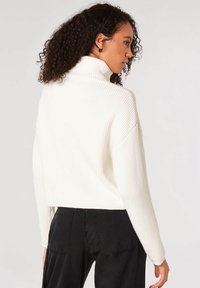 Maglione a collo alto bianco a coste con design corto e spalle scese, abbinato a pantaloni neri. Texture morbida e vestibilità rilassata.