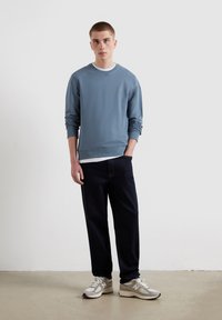 Felpa azzurro chiaro con colletto a girocollo e polsini a costine, indossata sopra una maglietta bianca. Pantaloni blu scuro e sneaker grigie completano il look.