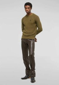 Uomo che indossa un maglione a maglia verde oliva, pantaloni in pelle marrone scuro con cinghie alle caviglie e stivali neri, in piedi di fronte a uno sfondo semplice.