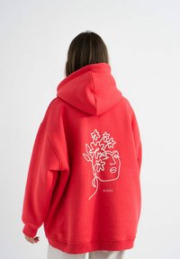Czerwona, oversize bluza z kapturem z dużym haftowanym wzorem twarzy i florystycznym motywem na plecach. Wykonana z miękkiego materiału, z kapturem i ściągaczami na mankietach.