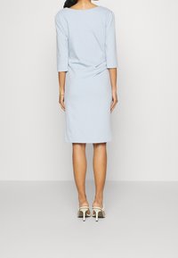 Vestido azul claro hasta la rodilla con mangas de tres cuartos, textura suave y detalle de espalda drapeada. Combinado con tacones de punta abierta en color crema.