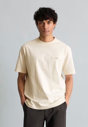 Mladý muž s kučeravými vlasmi, oblečený v krémovom tričku s krátkymi rukávmi a tmavými nohavicami, stojaci so založenými rukami v kapse na jednoduchom pozadí.