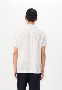 Camicia bianca a maniche corte con un design tessuto ad intreccio aperto, caratterizzato da motivi verticali. Con colletto, vestibilità ampia e orlo dritto.