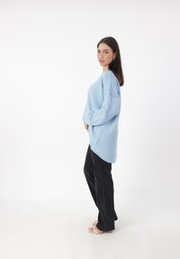 Mujer con un suéter oversized azul claro, pantalones negros y sandalias blancas, de pie de lado contra un fondo blanco plano.