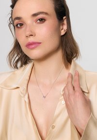 Srebrny naszyjnik z łańcuszka z okrągłym wisiorkiem z przezroczystym kamieniem, noszony na satynowej beżowej koszuli o miękkiej fakturze.