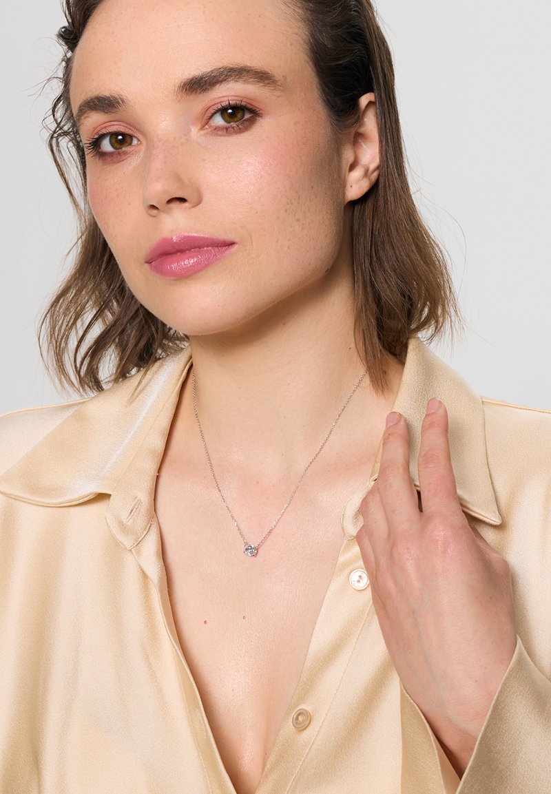 Srebrny naszyjnik z łańcuszka z okrągłym wisiorkiem z przezroczystym kamieniem, noszony na satynowej beżowej koszuli o miękkiej fakturze.