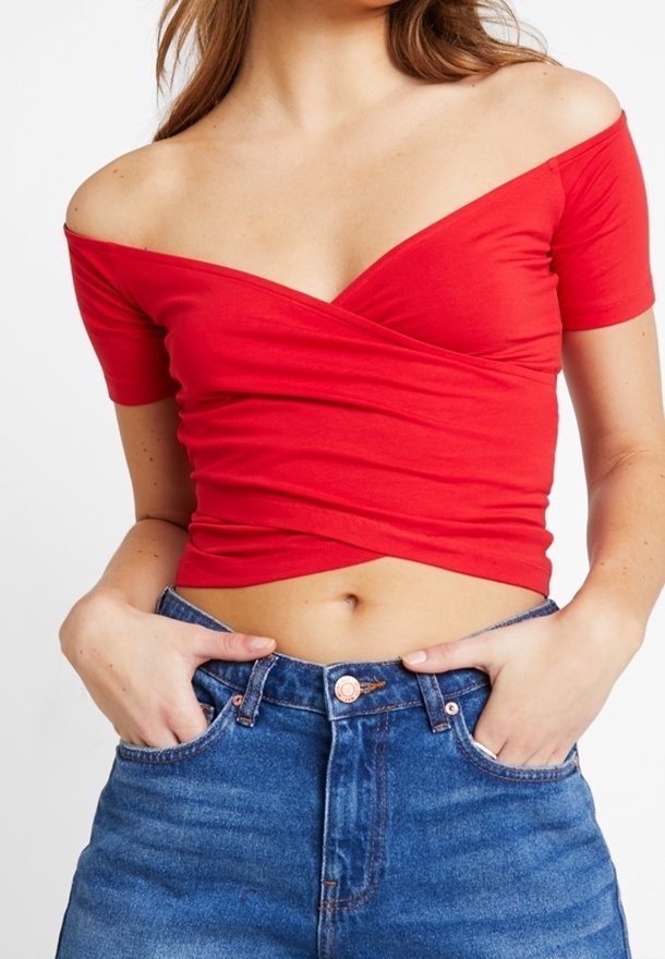 Kvinde iført en rød off-shoulder wrap crop top og blå højtaljede jeans med hænderne i lommerne mod en hvid baggrund.