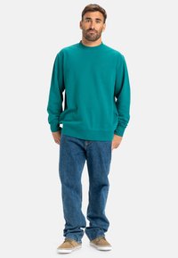 Teal sweatshirt med rund halsudskæring og ribstrikkede manchetter, parret med blå jeans og beige sko. Materiale i bomuldsblanding, minimal branding.