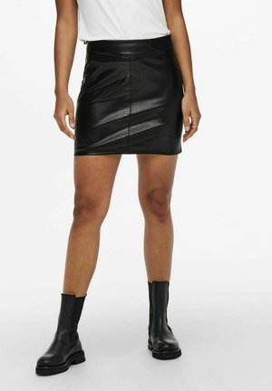 Femme portant une mini-jupe en similicuir noir et des bottines noires, debout devant un fond blanc.