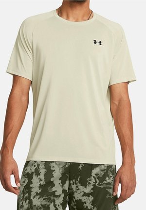 Homme portant un t-shirt beige à manches courtes Under Armour et un short de sport camouflage vert-gris, debout les bras détendus le long du corps.