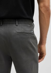 Grå byxor med en slät textur, utrustade med en bakficka och en liten "BOSS"-etikett. midjelinningen har bältesslingor.