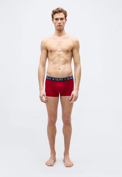 Νεαρός άνδρας όρθιος, ξυπόλυτος, φοράει κόκκι να μπόξερ με μαύρη ζώνη μέσης με το λογότυπο "Polo Ralph Lauren" σε απλό ανοιχτό γκρι φόντο.