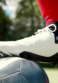 Scarpa da calcio bianca con accenti neri posizionata su un pallone da calcio nero. La scarpa ha una superficie testurizzata e lacci. Calzino rosso visibile sopra.