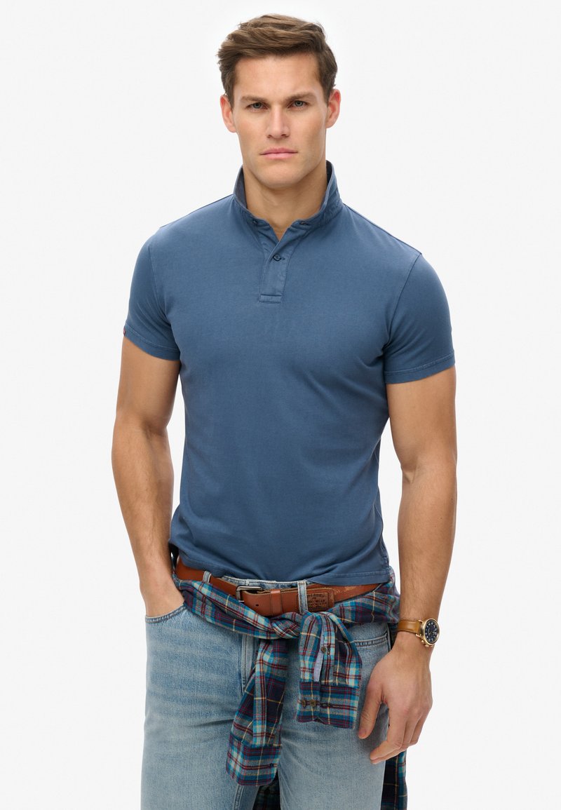Blauwe polo met korte mouwen en een button-down kraag. Gecombineerd met lichtgewassen jeans en een geruit shirt om de taille geknoopt. Bruine riem en horloge.