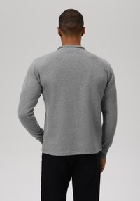 Camisa de manga larga gris hecha de tela suave, con un diseño sencillo y un dobladillo redondeado en la parte trasera. Sin logotipos ni patrones visibles.