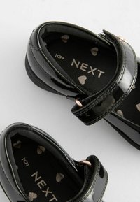 Schwarze Lacklederschuhe mit zwei verstellbaren Riemen, ausgestattet mit einer weichen schwarzen Innenausstattung mit silbernen Herzmustern und einer glatten Oberfläche.