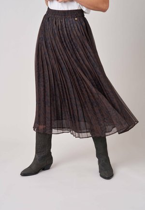 Femme portant une jupe midi plissée foncée avec un subtil imprimé animalier et des bottes noires brodées mi-mollet, debout devant un fond uni.