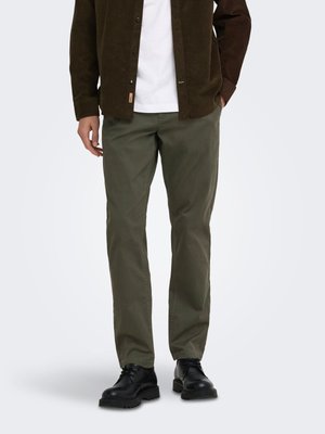 Homme portant un pantalon droit vert olive, des chaussures noires à lacets, un t-shirt blanc et une veste marron foncé boutonnée, debout les mains dans les poches.