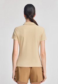 Beżowa koszula polo o miękkiej teksturze, z krótkim rękawem i klasycznym kołnierzykiem. W zestawieniu z brązowymi szortami, które prezentują swobodny design.