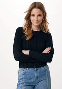 Černý žebrovaný kardigan s dlouhými rukávy, předními knoflíky a zvlněným výstřihem. V kombinaci s džínami s vysokým pasem v modré barvě.