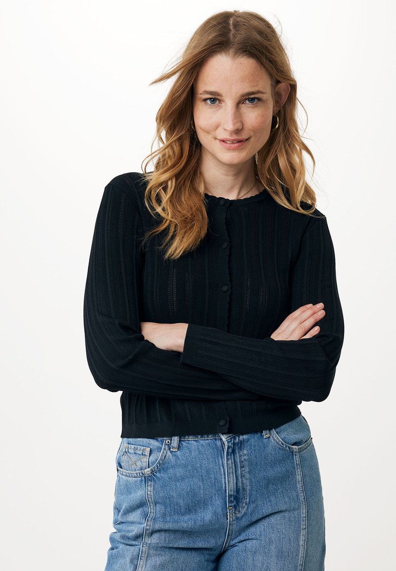 Černý žebrovaný kardigan s dlouhými rukávy, předními knoflíky a zvlněným výstřihem. V kombinaci s džínami s vysokým pasem v modré barvě.