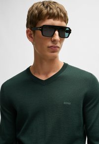 Młody mężczyzna w ciemnych prostokątnych okularach przeciwsłonecznych oraz ciemnozielonym swetrze z dekoltem w serek z logo "BOSS" na piersi na jednolitym tle.