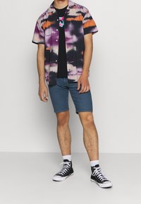 Kortärmad skjorta med knappar i svart, lila och orange batikmönster, bärs över en svart t-shirt, kombinerad med jeansshorts och svarta sneakers.