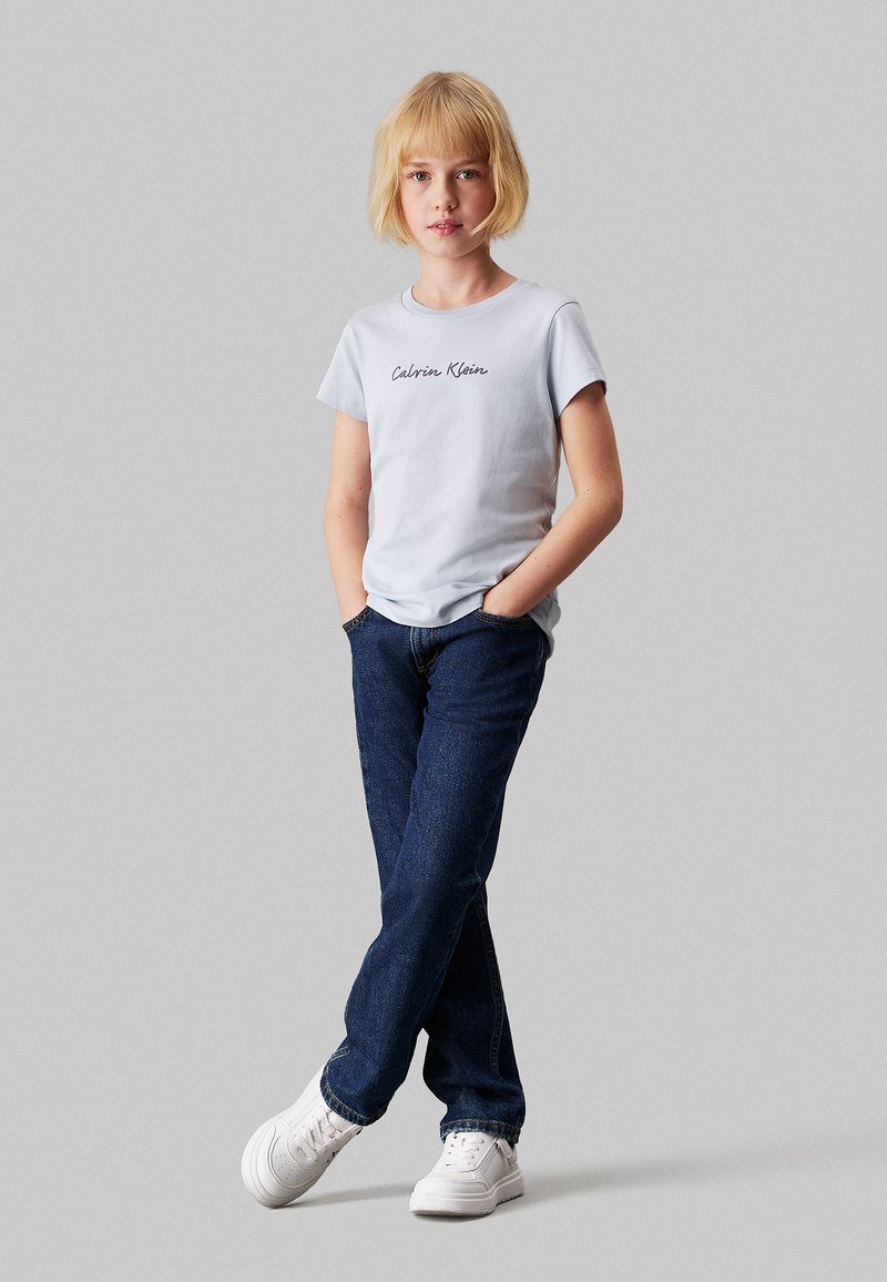 Camiseta de algodón de color azul claro con el logotipo de "Calvin Klein", combinada con jeans de mezclilla azul oscuro y zapatillas blancas, de pie con las manos en los bolsillos.