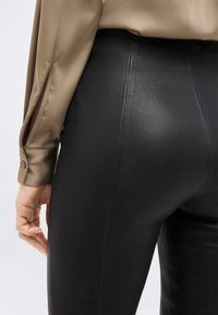 Κοντινό πλάνο ενός ατόμου που φοράει μαύρο παντελόνι από συνθετικό δέρμα και μπεζ σατέν μπλούζα με μανσέτα με κουμπιά και άνοιγμα στο μανίκι.