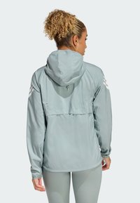 Himmelblaues Sportjackett mit Kapuze, ausgestattet mit einer Rückentasche, elastischen Bündchen und weißen Drei-Streifen-Akzenten auf den Schultern.