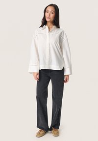 Witte blouse met geborduurde accenten en brede mouwen, gecombineerd met zwarte jeans en tan schoenen. Eenvoudig, casual ontwerp.