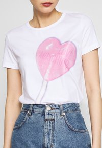 Bílý bavlněný tričko s velkým lesklým růžovým srdcovým lízátkem a textem "LOVE MOSCHINO", v kombinaci s vysokými džínovými šortkami.
