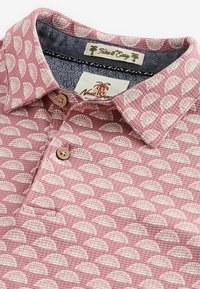 Polo en coton rose avec motif éventail blanc, doté d'un col, d'une patte à trois boutons, et d'une étiquette tissée avec un dessin de palmier.