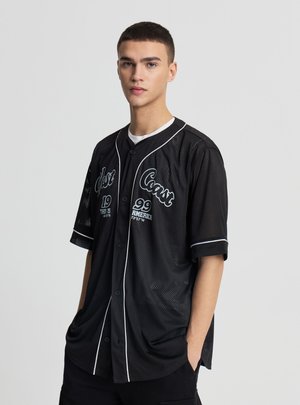 Giovane uomo con capelli corti che indossa una maglia da baseball nera con la scritta West Coast 99 America, in piedi con le mani in tasca davanti a uno sfondo uniforme.