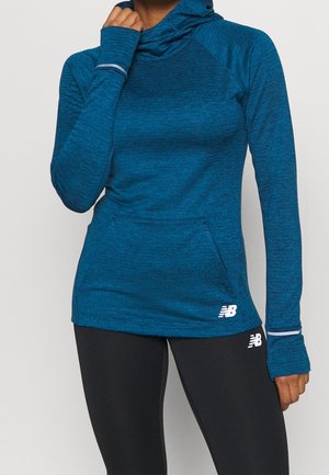 Personne portant un sweat à capuche bleu à manches longues avec des trous pour le pouce et des leggings noirs, les deux avec des logos New Balance blancs.