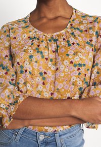 Frau mit verschränkten Armen, die eine gelbe Bluse mit lila, weißen und türkisfarbenen Blumen trägt, kombiniert mit hellblauen Lee-Jeans.