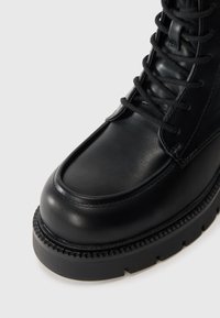 Black leather boot with a rounded toe, thick sole, and visible lacing. Features textured detailing around the sole edge and smooth finish.