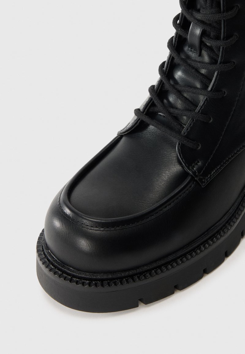 Stivale in pelle nera con punta tondeggiante, suola spessa e lacci visibili. Presenta dettagli testurizzati attorno al bordo della suola e una finitura liscia.