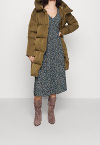 Oliivinvihreä puhvitakki, jossa on suuri huppu, tummakukkaistavarinen midi-mekko ja taupe-väriset löysät nilkkurit, joissa on paksut korot.