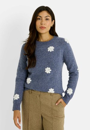 Suéter azul de punto con aplicaciones de flores blancas. Cuello redondo, mangas largas, ajuste ceñido y tela texturizada. Combinado con pantalones beige.