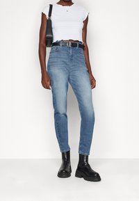 Frau trägt ein weißes, strukturiertes, kurzes Top, hochgeschnittene blaue Jeans mit schwarzem Gürtel, schwarze klobige Knöchelstiefel und schwarze Schultertasche.