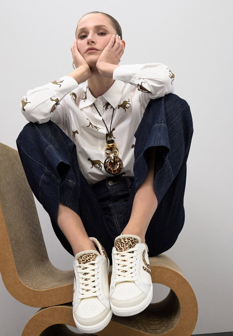 Camisa blanca con estampado de leopardo dorado, acompañada de pantalones vaqueros oscuros holgados y zapatillas blancas con un acento de estampado de leopardo en el talón.