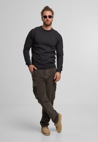 Pull en maille gris foncé, pantalon cargo olive avec poches, chaussures beiges à enfiler, et lunettes de soleil. Tenue décontractée avec des matériaux texturés.