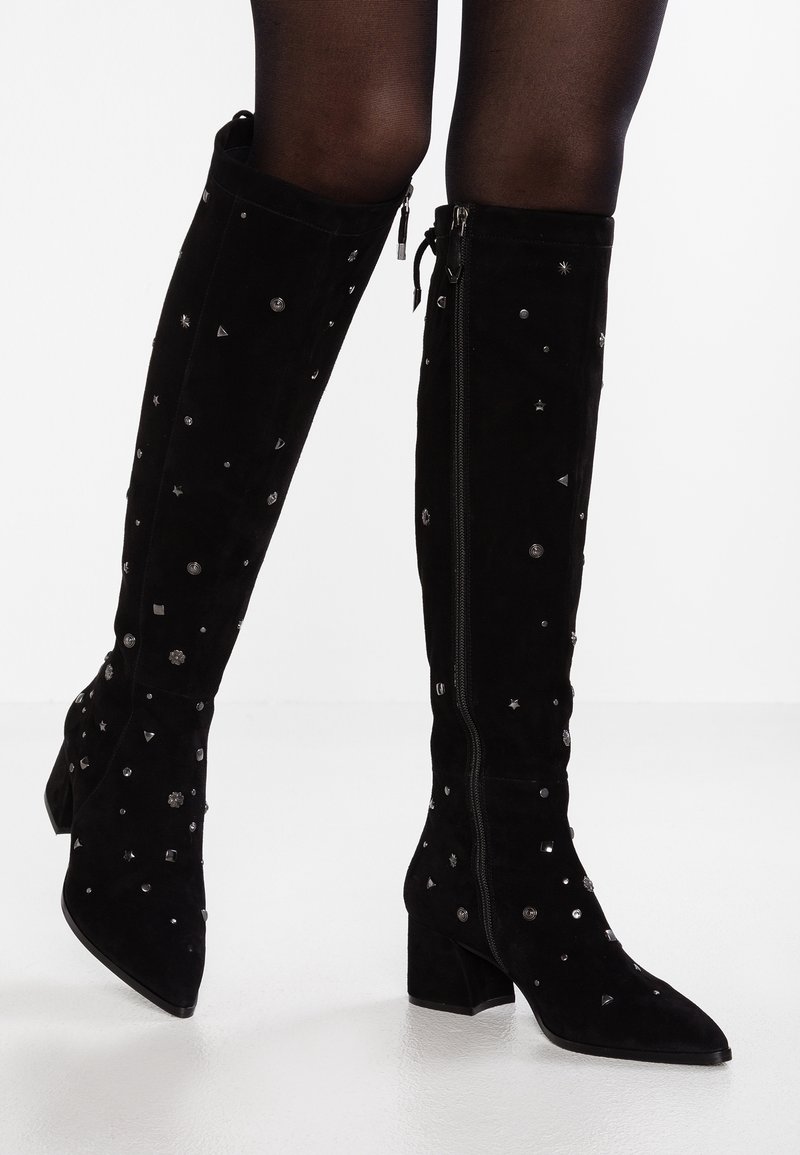 Mustat mokka polviin asti ulottuvat saappaat, joissa on paksut korot ja hopeiset niitit, yhdistettynä läpikuultaviin mustiin sukkahousuihin valkoisella lattia-taustalla.